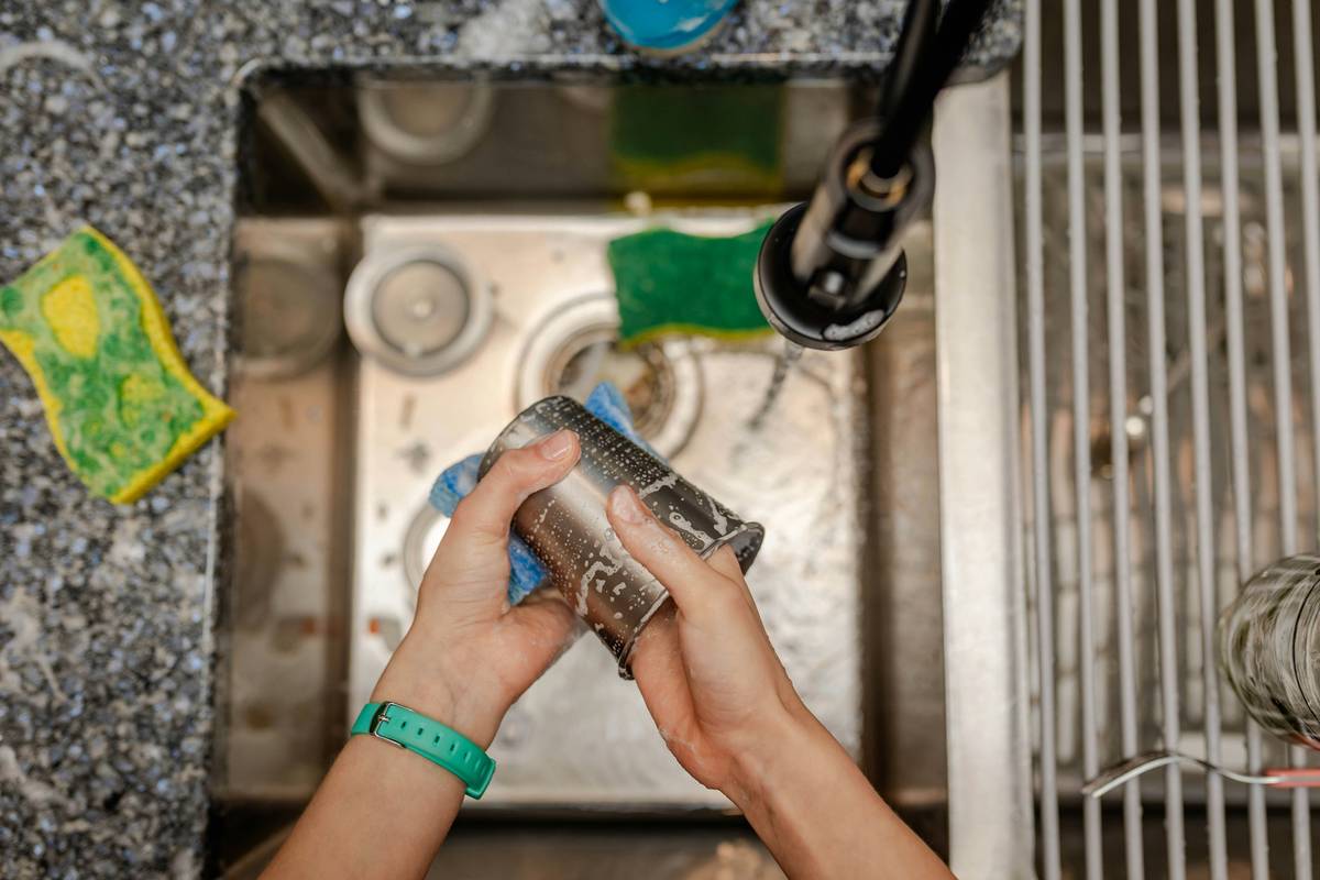 Close-up of stainless steel meat grinder auger, cutting blade, and plastic hopper arranged beside a dishwasher rack with 'meat grinder parts in dishwasher' warnings visible on the label